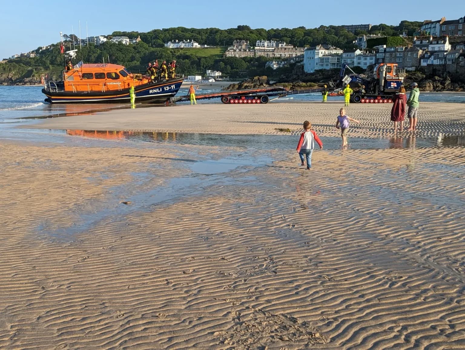A wide sandy beach at low tide with a lifeboat, a harbour town behind, and a few people far from the camera