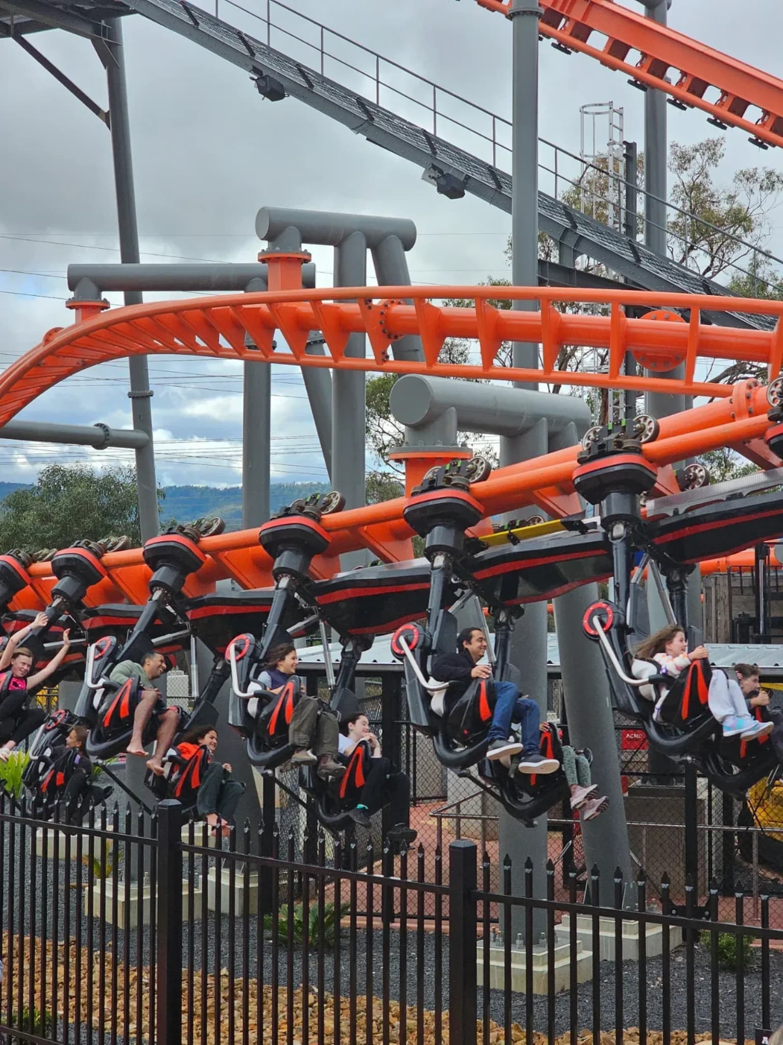 Looking up at an orange rollercoaster track with a train of riders hanging beneath it, railings in the foreground