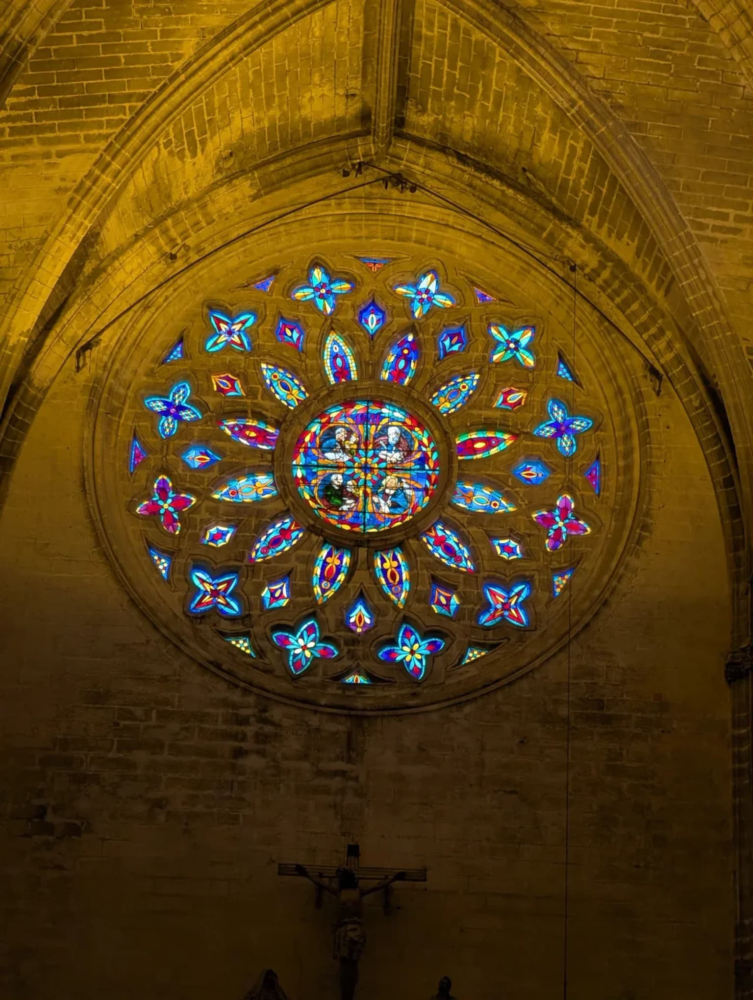 A round stained glass window set in a stone arch, lit from outside, with a small crucifix below it