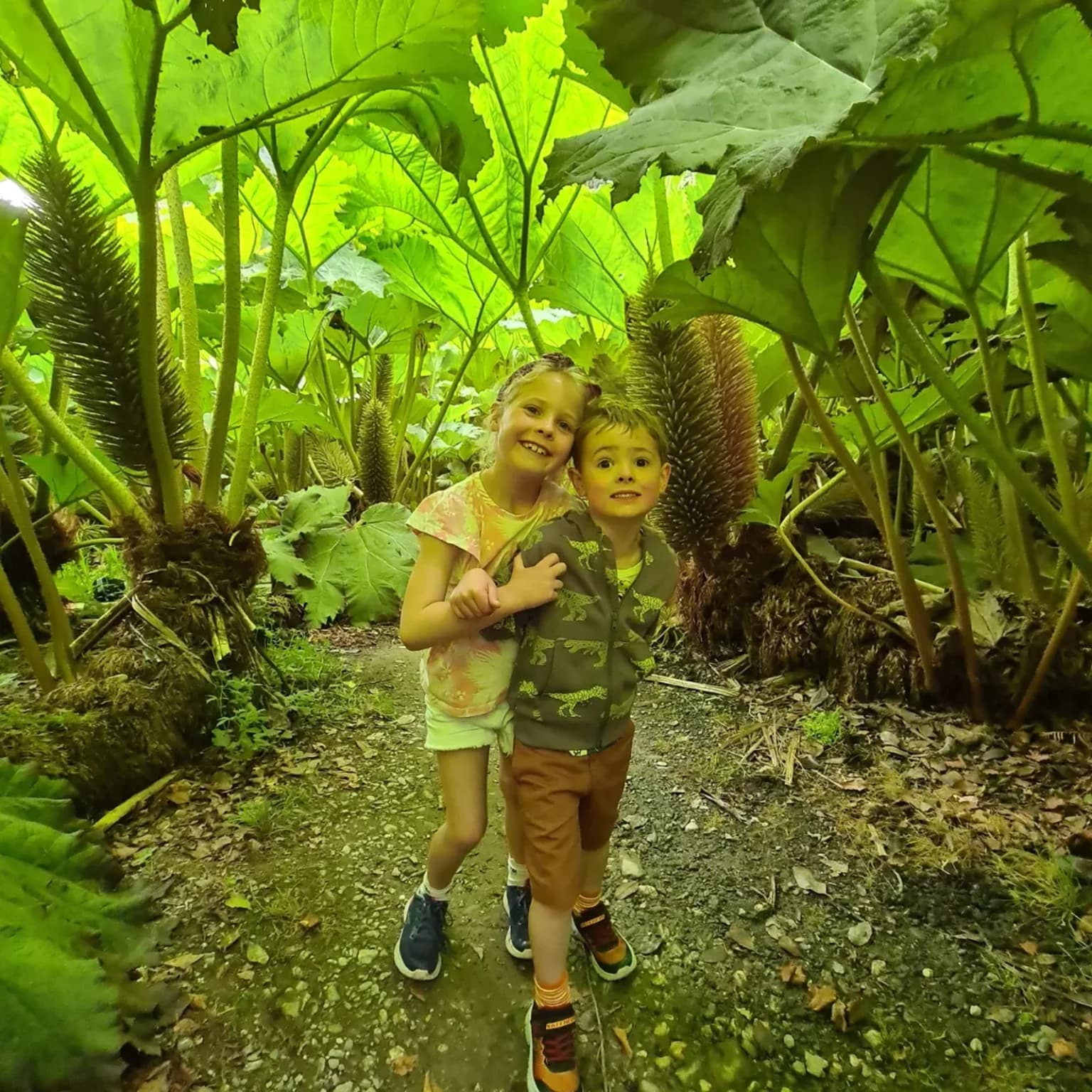 Two children standing close together on a garden path, arms round each other, with huge green leaves and tall flower spikes behind them